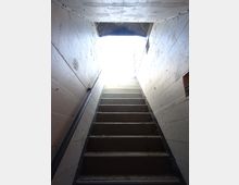 Scala in cemento all’interno di un passaggio stretto con pareti in calcestruzzo grezzo e corrimano metallico, che sale verso un’apertura esterna molto luminosa. Il soffitto basso e le superfici segnate evidenziano un ambiente tipo seminterrato o bunker.