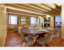 Ampio soggiorno-pranzo in un appartamento con pavimento in legno e soffitto con travi a vista, con tavolo rotondo apparecchiato e sedie moderne in primo piano. Sullo sfondo ci sono divani, librerie integrate e grandi finestre, con un passaggio che conduce a una camera da letto.