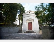 Piccola cappella in stile neoclassico con facciata bianca, frontone triangolare e portale ad arco con doppia porta in legno rosso. Davanti c’è un piazzale in pietra con gradini e muretti, circondato da alberi alti e vegetazione.