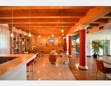 Interno di una sala ristorante o lounge con soffitto in travi di legno a vista, lampadine sospese e tavoli disposti su pavimento piastrellato lucido. A sinistra si vede un bancone e una libreria a parete, mentre a destra ci sono colonne rosse con capitelli bianchi, un corridoio e una pianta in vaso.