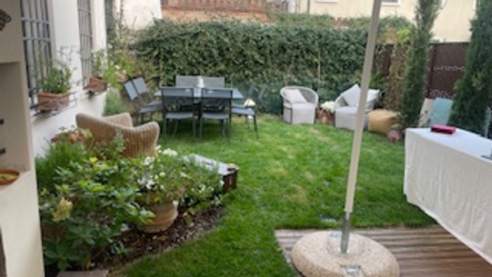 Giardino privato con prato curato, aiuole fiorite e zona pranzo con tavolo e sedie vicino a siepi e recinzioni. In primo piano una pedana in legno con ombrellone e un tavolo coperto da tovaglia bianca.