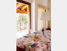 Sala da pranzo luminosa con tavolo apparecchiato, tovaglia a motivi marini, piatti bianchi e bicchieri, con lampada da tavolo e bottiglie al centro. Accanto a una grande porta-finestra che si apre su un balcone con ringhiera in legno e tettoia, affacciato sul verde esterno.