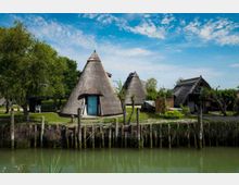 Capanni tradizionali con tetti di paglia a forma conica e una piccola casa in legno, situati su un prato alberato lungo un canale. In primo piano si vede la riva con pali di legno e acqua verde, sullo sfondo cielo azzurro parzialmente nuvoloso.