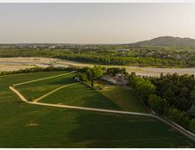 Veduta aerea di una campagna con campi coltivati e strade sterrate che convergono verso una villa storica con cortile, circondata da alberi. Sullo sfondo si vede un ampio letto di fiume ghiaioso, un’area urbana diffusa e colline basse.