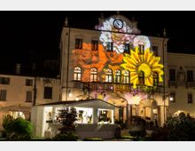 Facciata di un palazzo storico con portici e balconi, sormontato da un orologio centrale, ripresa di notte in una piazza illuminata. Sulla facciata sono proiettate grandi immagini colorate di fiori, tra cui un girasole e una rosa.
