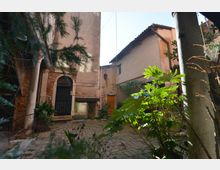 Cortile interno lastricato con vegetazione in primo piano, circondato da edifici in intonaco e mattoni a vista con tetti in coppi. Sul lato sinistro si vede un portale ad arco con grande porta scura e, sul fondo, un ingresso più piccolo sotto cavi e tubazioni esterne.