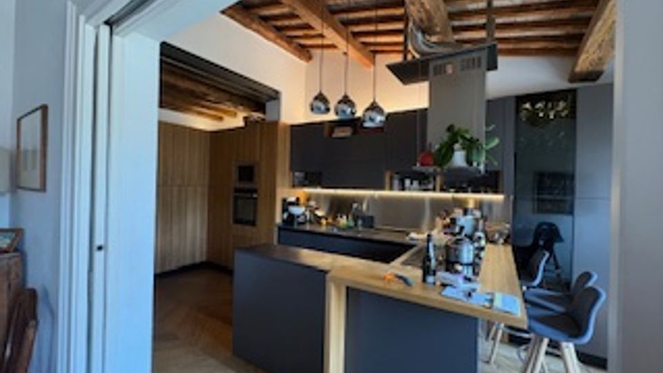 Cucina moderna open space con isola centrale e bancone in legno, mobili scuri e piano di lavoro attrezzato. Soffitto con travi a vista e luci a sospensione, con passaggio ad arco verso un’altra zona della casa.