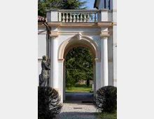 Arco d’ingresso in stile classico con colonne bianche e cornici beige, sormontato da una balaustra e da un piccolo volto decorativo al centro. Attraverso il passaggio si vede un vialetto che conduce a un’area verde con alberi, affiancato da siepi e una statua in pietra sul lato sinistro.