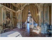 Interno di un salone storico riccamente decorato, con pareti e colonne ornate da stucchi e pannelli dipinti. Una grande tenda drappeggiata incornicia una porta che conduce a un’altra stanza illuminata, con pavimento in seminato e sedute lungo le pareti.