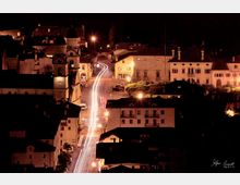 Veduta notturna di un centro storico con strada principale illuminata da lampioni e scie luminose delle auto, circondata da edifici con tetti scuri. In primo piano si distingue una torre dell’orologio e, sullo sfondo, un complesso con cupole e facciate storiche.