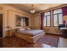 Camera da letto in stile classico con grande letto basso, pavimento in parquet e pareti con decorazioni a stucco e un grande quadro. Ampie finestre ad arco con tendaggi rossi illuminano la stanza, arredata con comodini, lampade e un mobile con televisore.