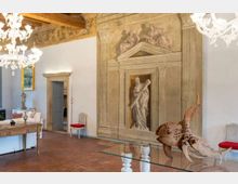 Interno di un salone storico con soffitto a travi in legno, grandi lampadari in vetro e pavimento in cotto. Una parete è decorata con un grande affresco architettonico con nicchia e figura classica; sono presenti arredi d’epoca e un tavolo in vetro con scultura in legno.