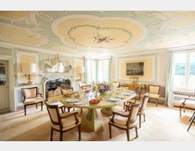 Sala da pranzo in stile classico con soffitto decorato a stucchi e affreschi, camino ornamentale e grande tavolo ovale apparecchiato circondato da sedie imbottite. Ampie finestre con tende chiare illuminano pareti pastello con boiserie e un dipinto, accanto a un mobile consolle dorato.