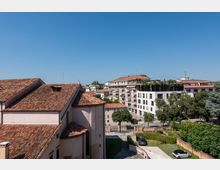 Vista urbana con tetti in coppi di un edificio storico in primo piano e palazzi residenziali moderni sullo sfondo. In basso si vedono alberi, un’area verde e una strada con parcheggi sotto un cielo sereno.