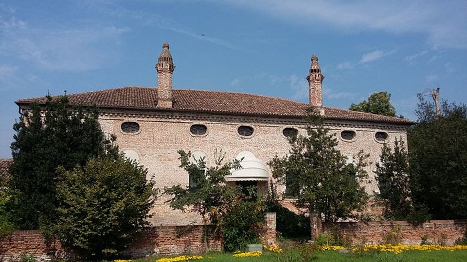 Edificio storico in mattoni con tetto in coppi e due alte canne fumarie, con una fila di piccole finestre ovali lungo la facciata. Davanti si vede un prato verde con fiori gialli, un muretto in mattoni e alberi e arbusti che parzialmente coprono l’ingresso con una piccola copertura arcuata.