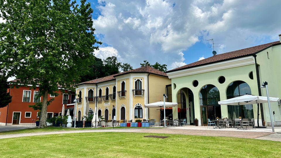 Complesso di edifici in stile villa con facciate colorate e tetti in coppi, affacciato su un prato verde con un grande albero. A destra si vede una struttura con ampie arcate e finestre circolari, con area esterna arredata con tavoli e ombrelloni.