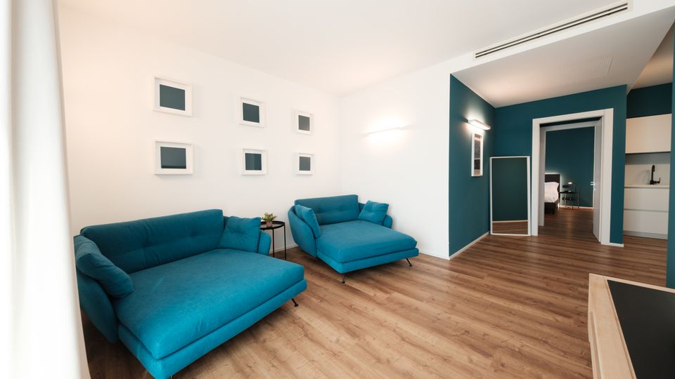 Soggiorno moderno con pavimento in legno, pareti bianche e una parete d’accento color petrolio, arredato con due divani turchesi e piccoli quadri minimalisti. Sul fondo si vede un corridoio con specchio a figura intera che conduce a una camera e a un angolo cucina.