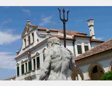 Statua in pietra vista da dietro con un tridente in primo piano, davanti a un edificio storico intonacato di bianco con tetto in tegole, finestre con scuri verdi e porticato ad archi, sotto un cielo azzurro.