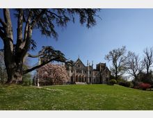 Villa storica in stile neogotico con archi e torrette, vista da un ampio prato verde in un parco. Grandi alberi incorniciano la scena e un albero in fiore si trova davanti all’edificio sotto un cielo sereno.