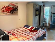Camera da letto con letto matrimoniale coperto da piumone a quadri e cuscini, pareti chiare con quadro di un’auto rossa. Sullo sfondo ci sono un armadio, una porta e una portafinestra che dà su un’area esterna, con pavimento chiaro e alcuni oggetti sparsi.