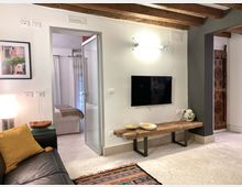 Interno di un soggiorno moderno con travi in legno a vista, pareti chiare e pavimento in pietra chiara. Sulla parete c’è una TV a schermo piatto sopra una panca in legno rustico, con porta aperta che conduce a una camera da letto.