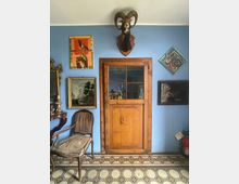 Interno di una stanza con parete azzurra e porta in legno con finestra a riquadri, circondata da quadri e cornici. Pavimento in piastrelle geometriche e arredi d’epoca, con una sedia imbottita e uno specchio su mobile lungo il lato sinistro.