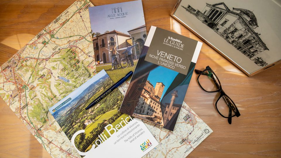 Piano in legno con mappe cartacee del Veneto e materiali turistici, tra cui guide e brochure, disposti a ventaglio. Sono visibili anche una penna, un paio di occhiali e una stampa incorniciata con disegno architettonico.