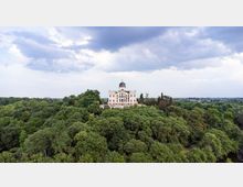 Veduta aerea di una villa storica con facciata chiara e cupola centrale, situata sulla sommità di una collina e circondata da un fitto bosco. Sullo sfondo si vede la pianura con campi e edifici sparsi sotto un cielo nuvoloso.