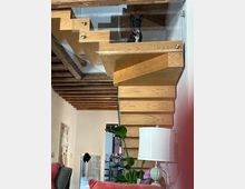 Interno domestico con scala moderna in legno a sbalzo e ringhiera in vetro, sotto un soffitto con travi a vista. Accanto alla scala si vedono una pianta in vaso, un tavolino con lampada e una parete decorata con quadri.