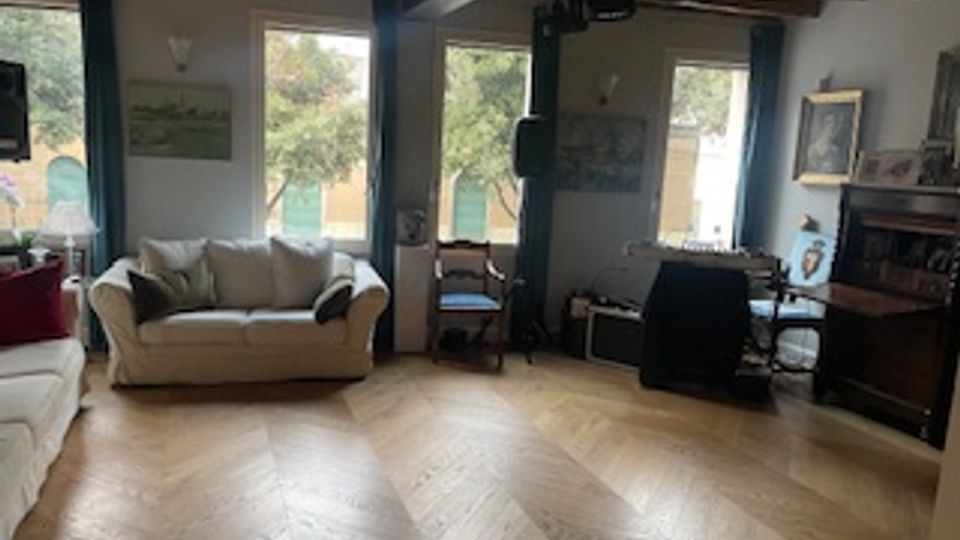 Interno di un soggiorno rustico con travi in legno a vista, pavimento in parquet a spina di pesce e grandi finestre che illuminano la stanza. Sono presenti divani, un tavolo con sedie, poltrone e un mobile scuro lungo la parete con quadri e oggetti decorativi.