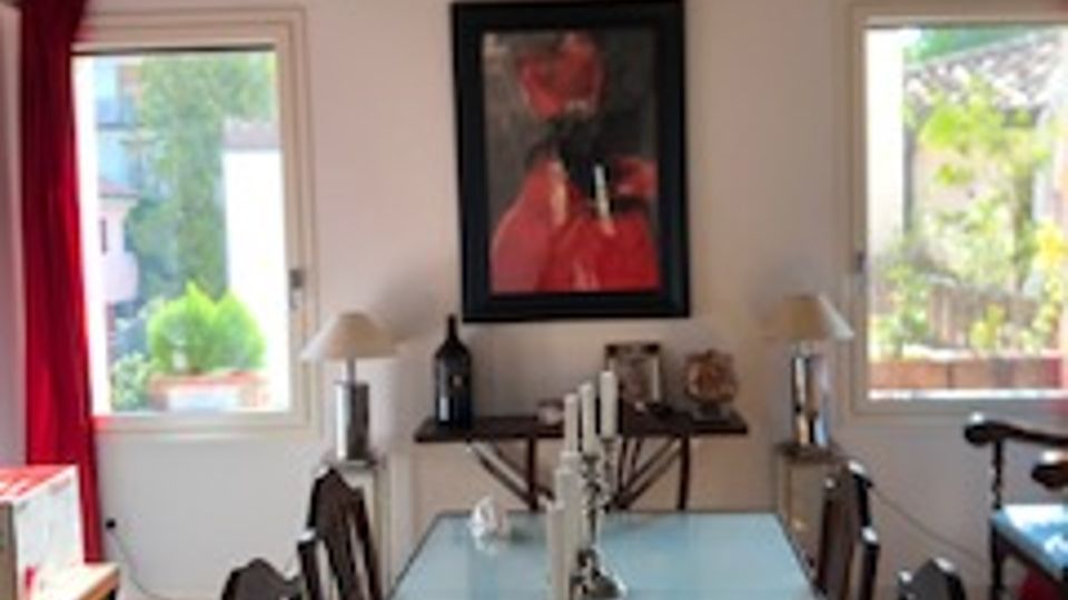 Sala da pranzo in stile rustico con lungo tavolo rettangolare e sedie scure, lampadario decorativo e soffitto con travi in legno a vista. Due finestre con tende rosse illuminano la stanza, con credenza sul fondo e un quadro appeso alla parete.