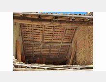 Sottotetto di un edificio rurale con struttura del tetto in travi di legno e file di coppi in terracotta, visto dall’interno. Le pareti sono in muratura grezza e lungo il bordo inferiore si notano assi e materiali accatastati.