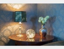 Angolo di soggiorno con tavolino rotondo in legno, lampada da tavolo con paralume verde e sfera luminosa decorativa. Sulla destra un vaso metallico con fiori chiari, davanti a parete con carta da parati damascata.