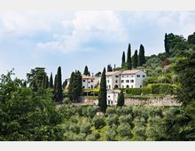 Complesso di edifici rurali in stile villa su una collina, con facciate chiare, tetti in coppi e una piccola cappella. Intorno si vedono cipressi alti, uliveti terrazzati e un muro in pietra, sotto un cielo parzialmente nuvoloso.