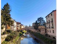 Veduta di un canale cittadino con un ponte in pietra ad arco al centro e case a più piani dai colori chiari affacciate sulle rive. Vegetazione lungo gli argini e alberi alti incorniciano la scena sotto un cielo sereno.