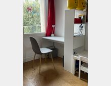 Angolo studio in una stanza luminosa con finestra, tende rosse e pavimento in parquet, composto da scrivania bianca con sedia grigia e una lampada da tavolo. Accanto c’è una libreria bianca con ripiani e un piccolo tavolino con sedie, con decorazioni e giocattoli in stile LEGO sulla parte superiore.