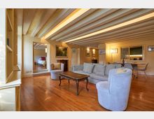 Ampio soggiorno open space con pavimento in legno e soffitto con travi a vista verniciate di bianco, arredato con divano, poltrone e tavolino. Sul fondo si vedono la zona pranzo con cucina a vista e, a sinistra, un’apertura che conduce a una camera da letto rialzata di pochi gradini.