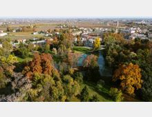 Vista aerea di un ampio parco con alberi dai colori autunnali che circondano un laghetto. Sullo sfondo si vedono edifici e una chiesa con alto campanile, con campi agricoli e area urbana all’orizzonte.