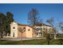 Piccola chiesa con facciata neoclassica e campanile a vela, affiancata da edifici rurali intonacati e tetti in coppi. Davanti si estende un prato con alberi spogli e un vialetto, in un contesto di campagna sotto cielo sereno.