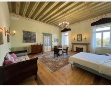 Ampia camera da letto in stile classico con soffitto a travi dipinte, lampadario centrale e pavimento in legno con grande tappeto decorato. Arredi in legno, letto matrimoniale, divanetto, tavolino con sedie, camino e grandi finestre con tende bianche affacciate sull’esterno.