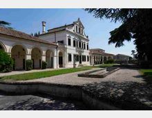 Cortile esterno di una villa storica con facciata chiara e finestre con scuri, affiancata da un porticato ad archi e da edifici annessi. In primo piano un’area ghiaiosa con vasche rettangolari basse e prati, circondata da alberi.