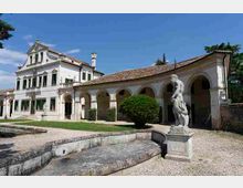 Cortile di una villa storica con facciata intonacata chiara e finestre con persiane verdi, affiancata da un lungo porticato ad archi e tetto in coppi. In primo piano si vede una vasca in pietra e una statua su piedistallo, con ghiaia, prato e alberi sullo sfondo.