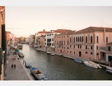 Veduta di un canale urbano con edifici storici dai tetti in tegole e facciate in intonaco su entrambe le sponde. Lungo le rive ci sono una passeggiata con tavolini e ombrelloni, barche ormeggiate e un ponte sullo sfondo.