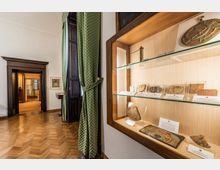 Interno di museo con pavimento in parquet a spina di pesce, alte porte in legno scuro e tenda verde accanto a una finestra. A destra, una vetrina a parete con ripiani in vetro espone strumenti e manufatti antichi con cartellini descrittivi.