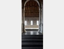 Interno di una sala storica con grandi colonne in pietra e arco centrale, pavimento in lastre scure e gradini che conducono a una piattaforma rialzata. Sul fondo si vedono tre alte finestre ad arco e pareti affrescate, sotto un soffitto a cassettoni in legno.