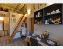 Interno di un piccolo appartamento mansardato con soffitto in travi di legno, scala in legno che porta a un soppalco e angolo studio sotto la scala. A destra ci sono mensole a muro con stoviglie e elettrodomestici e un tavolo da pranzo apparecchiato, con quadri alle pareti e illuminazione a parete.