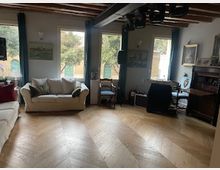 Interno di un soggiorno rustico con travi in legno a vista, pavimento in parquet a spina di pesce e grandi finestre che illuminano la stanza. Sono presenti divani, un tavolo con sedie, poltrone e un mobile scuro lungo la parete con quadri e oggetti decorativi.