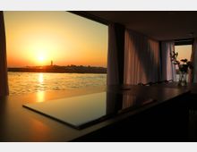 Interno moderno con grande finestra panoramica affacciata su un tratto d’acqua al tramonto, con riflessi dorati e profilo di edifici all’orizzonte. In primo piano un tavolo scuro lucido; ai lati tende bianche e un vaso con fiori vicino alla finestra.