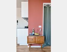 Interno domestico con parete color terracotta e pavimento in legno, con un piccolo mobile in legno a due cassetti appoggiato al muro. A sinistra si intravede una cucina moderna bianca con cappa, mentre a destra una porta con tenda verde scuro.