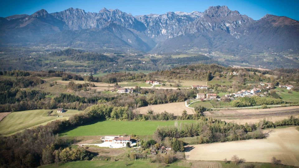 Veduta panoramica di una valle rurale con campi coltivati, boschetti e piccoli borghi sparsi, attraversata da strade di campagna. Sullo sfondo si alza una catena montuosa rocciosa sotto un cielo limpido.